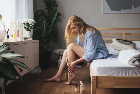 Woman sits on edge of bed and applies body oil to legs