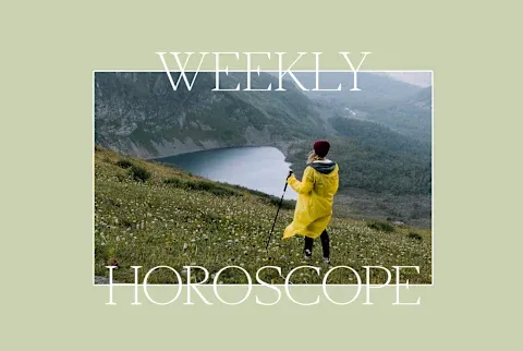 Woman in yellow rain jacket with a hiking stick stands on a hillside overlooking a vista with a lake