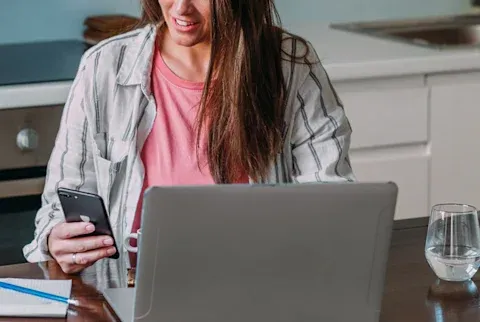 Brunette woman wearing pink shirt with sitting in front of a computer looking at her phone