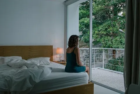 Woman in a nightgown sitting on bed facing window on a dark early morning